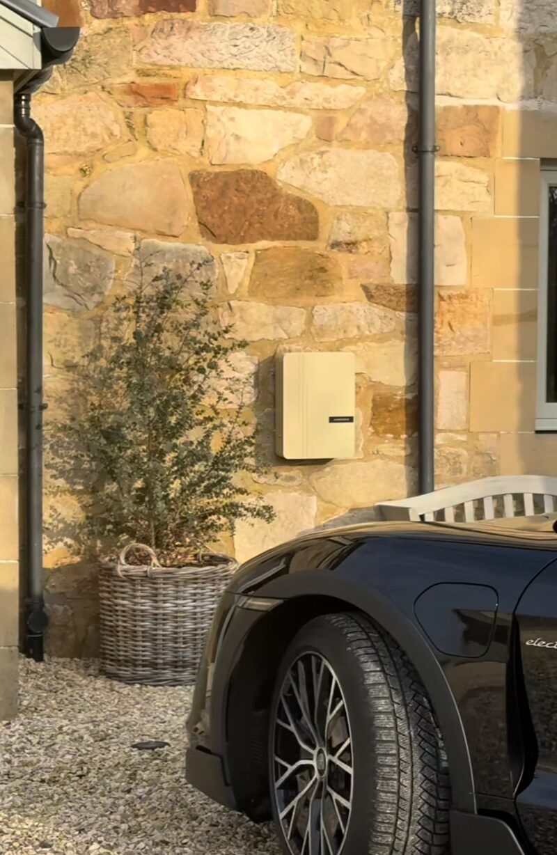 A cream Andersen A3 EV charger mounted on a stone wall beside a parked black electric car