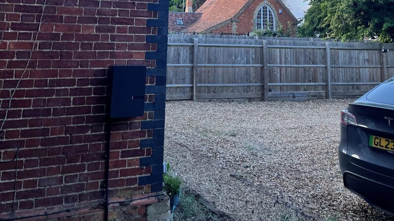 A black Andersen A3 EV charge point mounted on a brick wall beside a gravel driveway with a Tesla parked nearby