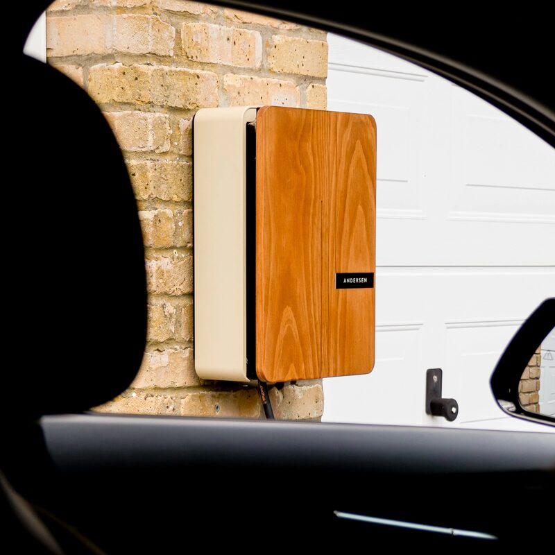 An Andersen EV home electric car charger with a wood-effect front panel mounted on a brick wall beside a garage door, seen through a car window