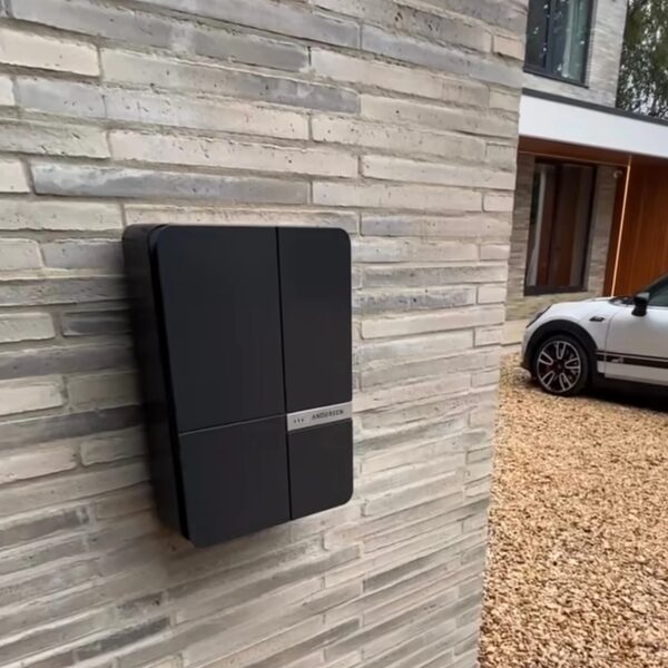 A black Andersen EV wall-mounted electric car charge point installed on a brick wall outside a modern house with a car parked on the gravel driveway