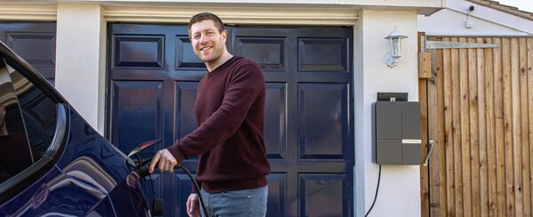 A man plugging a charging cable into an electric car on a driveway beside a wall-mounted Andersen EV charger