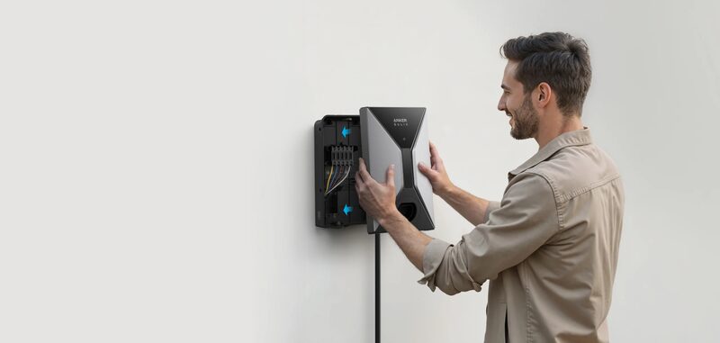 A man mounting an Anker SOLIX wall-mounted EV charger onto a white wall, with the back plate and wiring visible behind it