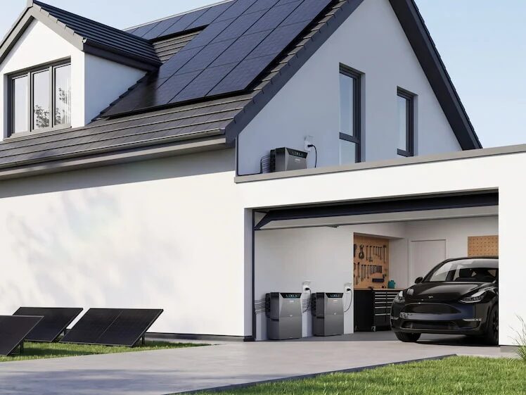A modern white house with rooftop solar panels, ground-mounted panels, Anker battery units in the garage, and an electric car charging