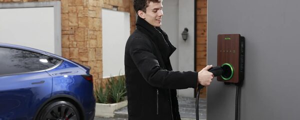 A man plugging a charging cable into a wall-mounted Autel EV charger outside a house, with a blue electric car parked nearby