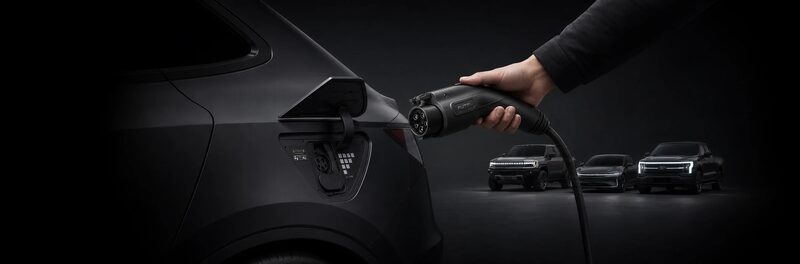A hand holding an EV charging connector plugged into a car's charging port, with other electric vehicles in the dark background