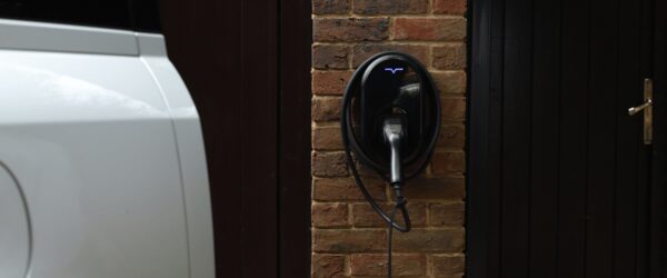 A black wall-mounted EV charger with a blue logo and coiled cable installed on a brick wall beside a white car