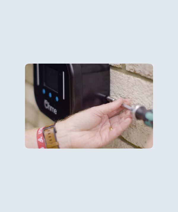 A person using a screwdriver to fix a black Ohme Home Pro EV charger to a brick wall