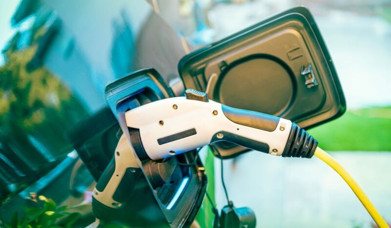 A close-up of an EV charging plug connected to a car's charging port with a yellow cable