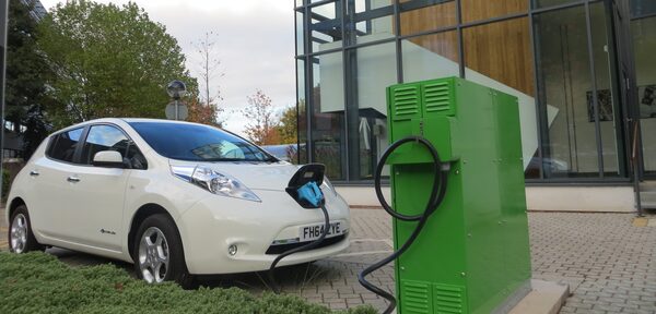 A white Nissan Leaf electric car plugged into a large green vehicle-to-grid charging unit outside a modern glass building