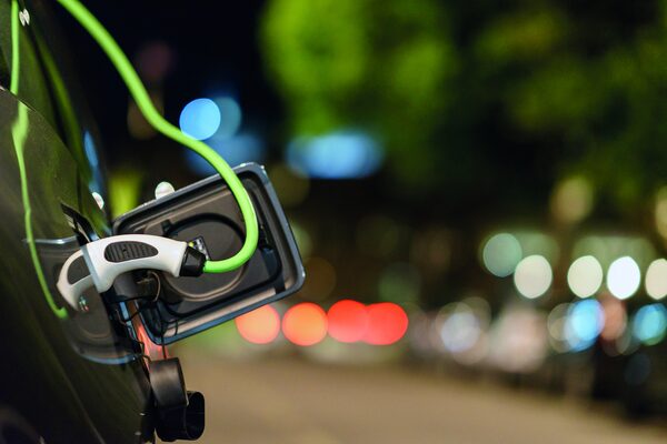 A green charging cable plugged into the charging port of an electric car at night with blurred city lights in the background