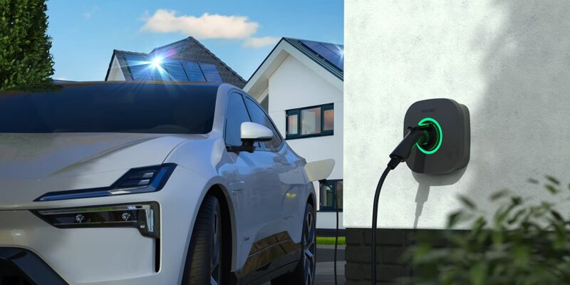A white electric car charging from a wall-mounted Rolec EV charge point outside a home with rooftop solar panels