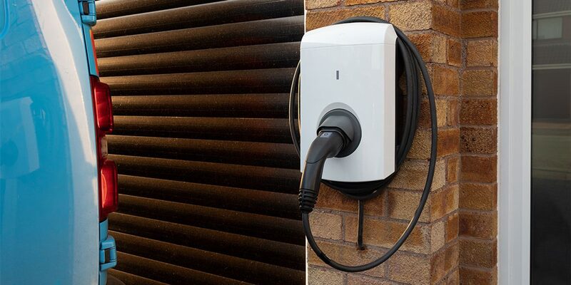 A white wall-mounted electric car chargepoint with a coiled cable and plug on a brick house wall next to a blue car