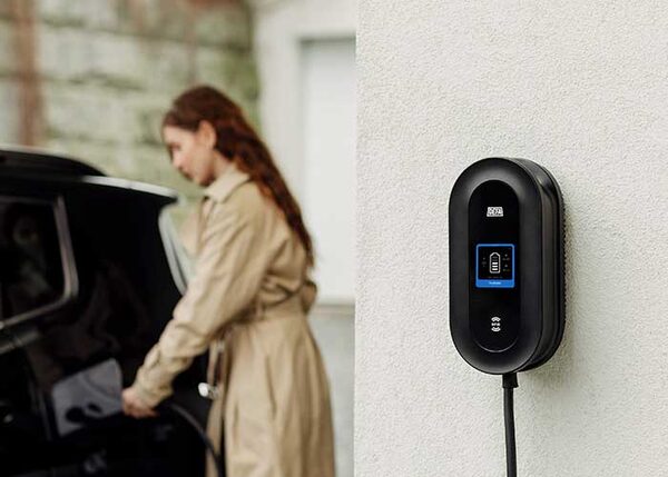 A black DEFA wall-mounted EV home charger on a white exterior wall with a woman charging a car in the background