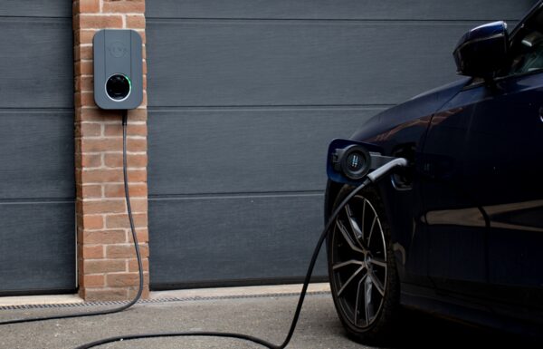 A Veva wall-mounted EV charge point on a brick pillar charging a dark blue electric car outside a garage