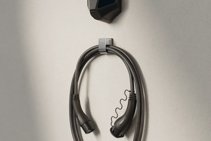 A black Easee wall-mounted EV charger with its coiled charging cable hung on a holder against a plain wall