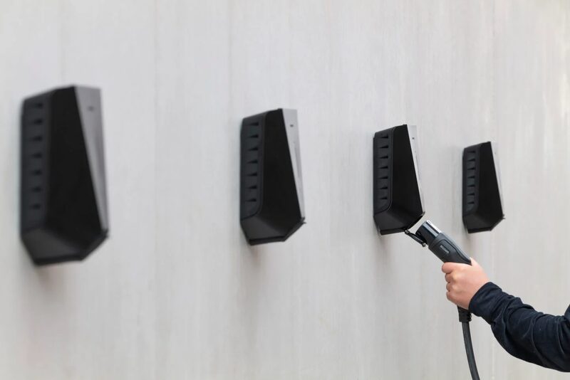 A row of four black Easee EV wall chargers mounted on a light wall, with a hand plugging a charging cable into one