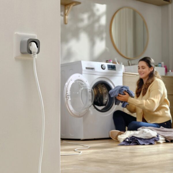 A smart plug with a washing machine cable plugged into a wall socket while a woman loads laundry in the background