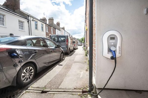 A grey electric car charging from a Pod Point wall charger on a house wall beside a residential street