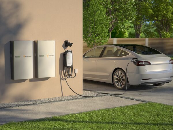 An Enphase home EV charger mounted on a house wall charging a white electric car in a driveway, with two Enphase battery units beside it