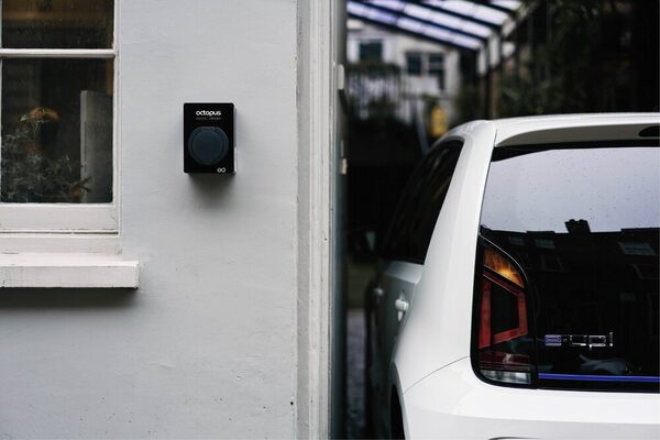 A black Octopus-branded EO home EV charge point mounted on a white house wall beside a parked white Volkswagen e-up! electric car