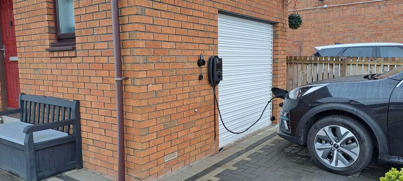 A wall-mounted EV charge point on a brick house charging a dark electric car on the driveway