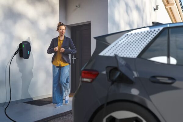 A wall-mounted EV charge point outside a home charging a dark electric car, with a woman walking out of the doorway