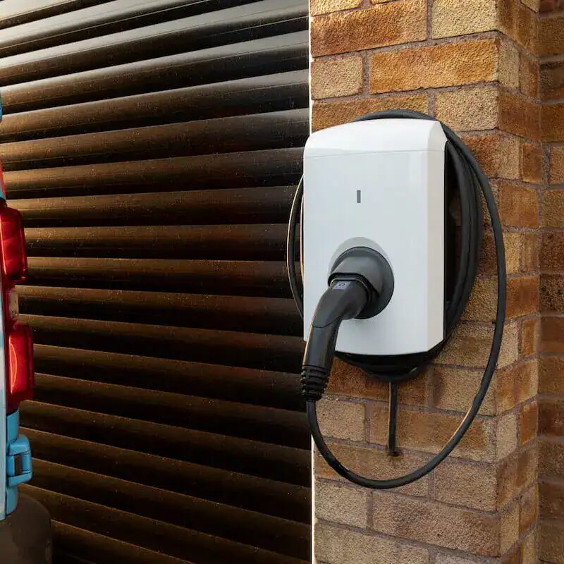 A white wall-mounted EV charge point with coiled cable and plug on a brick house wall next to a garage door