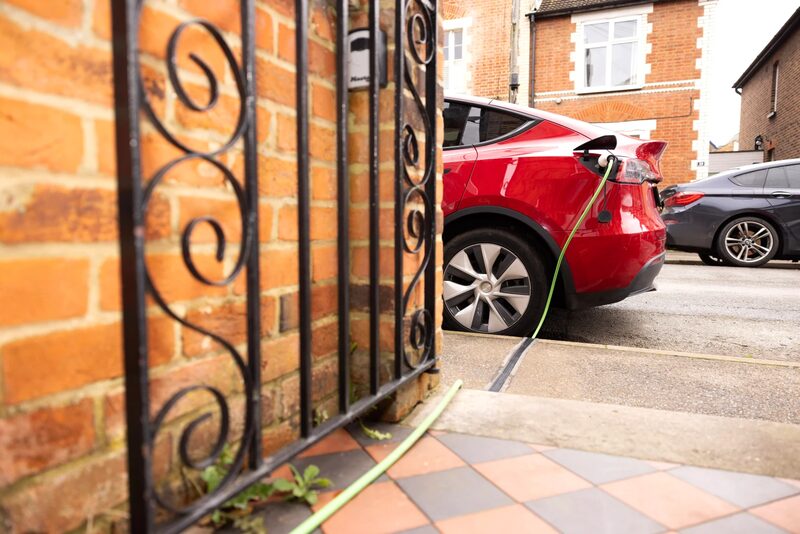 A red electric SUV parked on a residential street charging via a green cable running through a pavement gully (Kerbo Charge style cross-pavement charging)