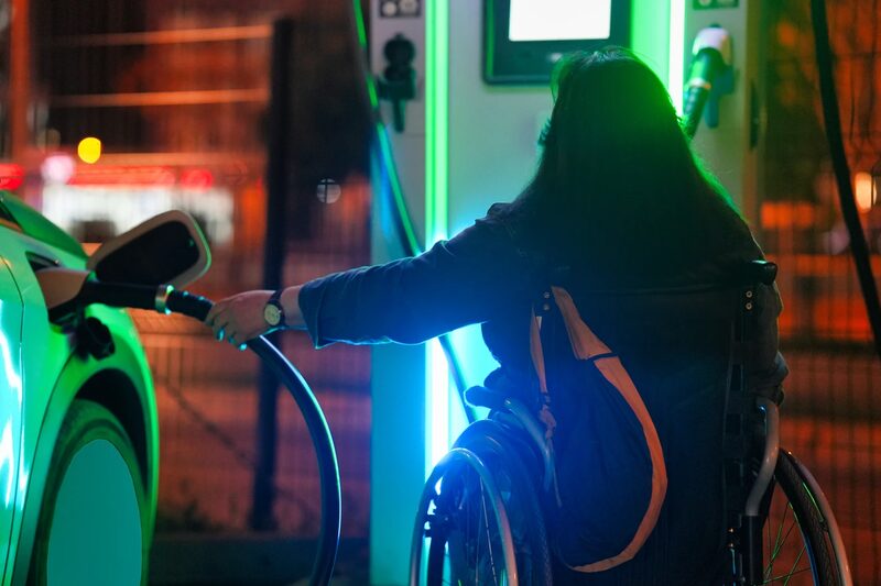 A woman in a wheelchair plugging a charging cable into an electric car at a public charge point at night