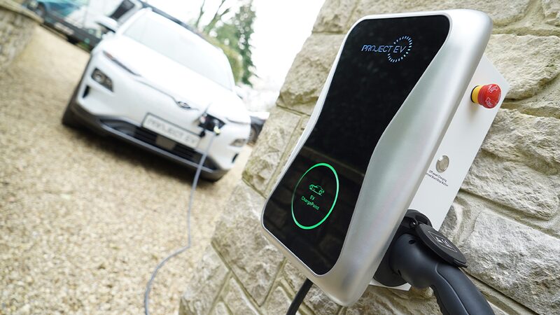 A white Project EV wall-mounted home EV charger on a stone wall with a charging cable plugged in and an electric car charging in the background