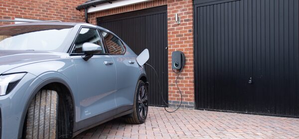A grey electric car charging from a wall-mounted EV charger on a brick house driveway