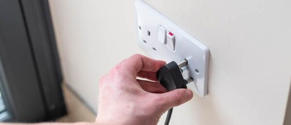 A hand plugging a black three-pin plug into a white UK wall socket