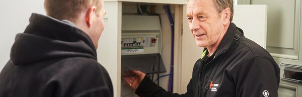 An electrician in a branded fleece showing a homeowner a consumer unit (fuse box) in a hallway