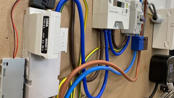 A close-up of an electricity meter and incoming supply fuse with coloured cables on a wooden board