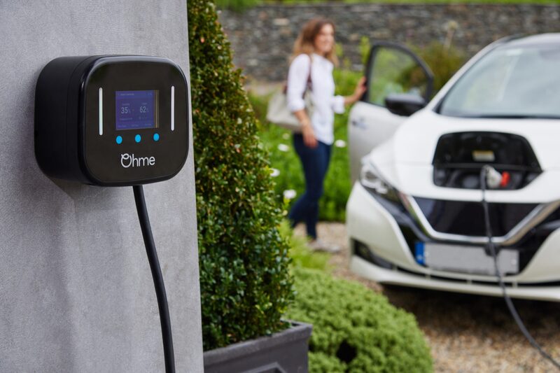 An Ohme wall-mounted EV charger on a house wall with a cable plugged into a white electric car in the driveway