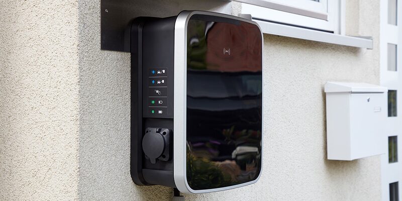 A black and silver wall-mounted electric car chargepoint with status lights and a socket cover, fitted to the exterior wall of a house next to a white postbox