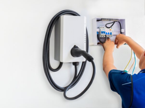 An electrician wiring a wall-mounted EV charger next to a small consumer unit on a white wall