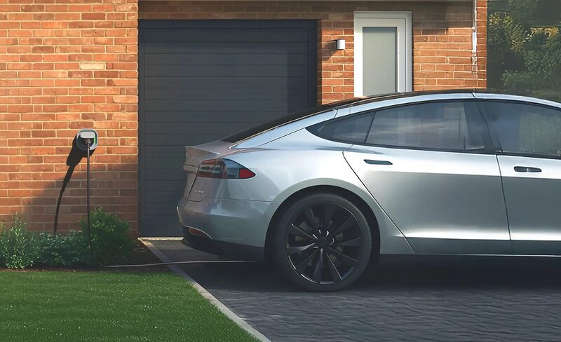 A silver electric car parked on a driveway charging from a wall-mounted EV charger outside a brick house