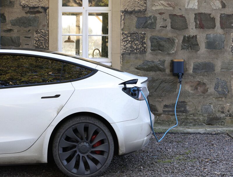A white electric car charging from a wall-mounted EV charger on a stone house wall
