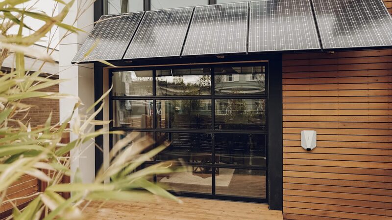 A white Easee EV charger mounted on a timber-clad house wall beneath a canopy of solar panels above a glass garage door