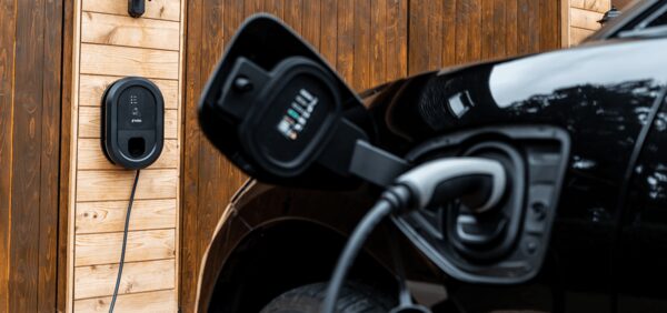 A black Ratio EV home wall charger mounted on a wooden wall with a cable plugged into a black electric car's charge port