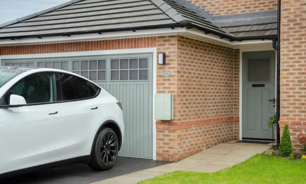 A white electric car parked on a driveway next to a wall-mounted EV charger on a brick house