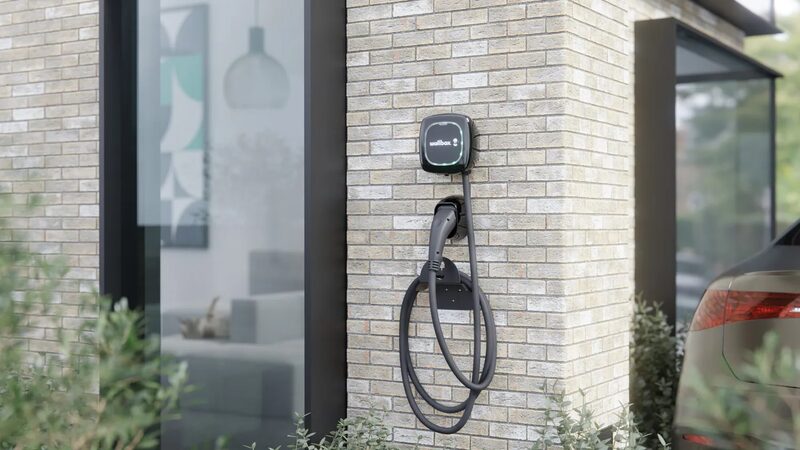 A Wallbox Pulsar Max EV charger mounted on a brick house wall with coiled cable and a car parked nearby