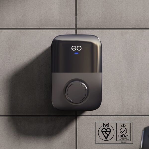 A black EO wall-mounted EV charge point with a Type 2 socket on a grey tiled wall, with BSI and UKAS certification marks in the corner