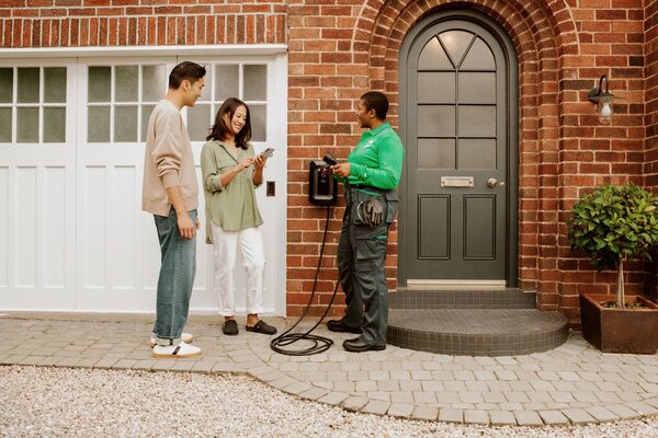 An installer in a green uniform demonstrating a wall-mounted EV charger to a couple outside a brick house