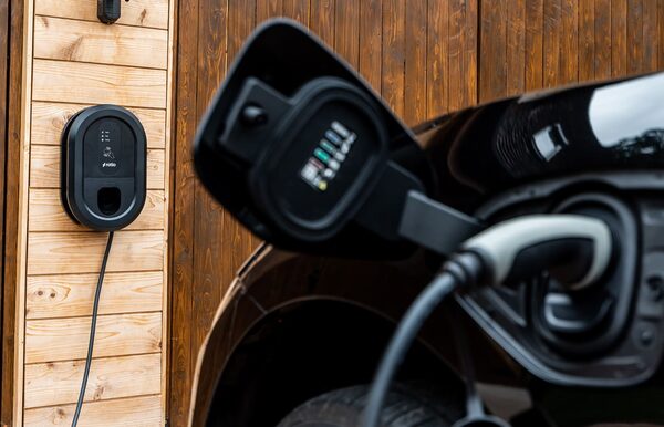 A black Ratio EV wall charger mounted on a wooden wall with a charging cable plugged into a car's charge port