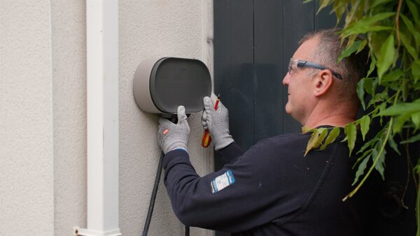 An engineer in gloves and safety glasses installing a wall-mounted EV charger on an exterior house wall