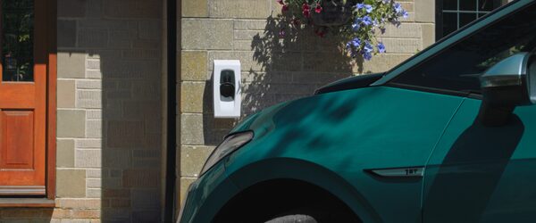 A white wall-mounted home EV charger on a brick house wall next to a parked teal car