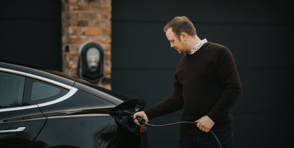 A man plugging a charging cable into a black electric car outside a home with a wall-mounted EV charger