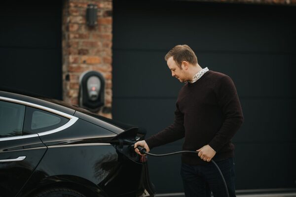 A man plugging a charging cable into a black electric car outside a home with a wall-mounted EV charger